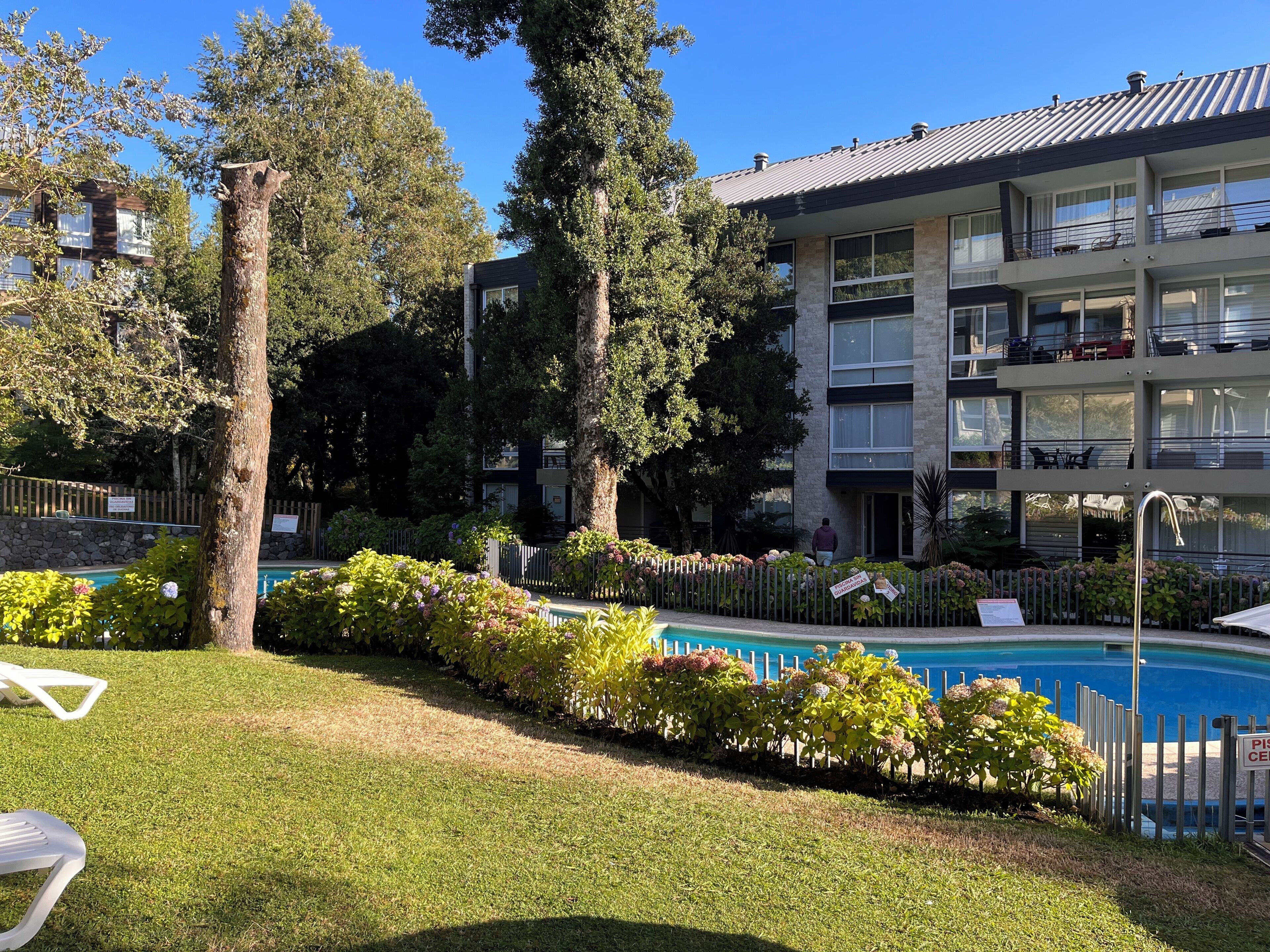 A pool area with a fence, lounge chairs, and a multi-story building with balconies.