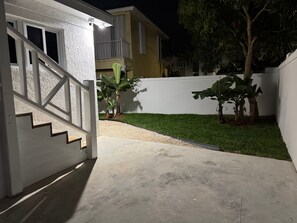 Un quartier résidentiel avec un chemin en béton menant à une maison blanche dotée d’un balcon. On y trouve deux bananiers et une petite pelouse.