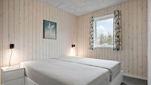 A bedroom with a bed, a nightstand, a wall-mounted lamp, a window with floral curtains, and a painting of a bird.