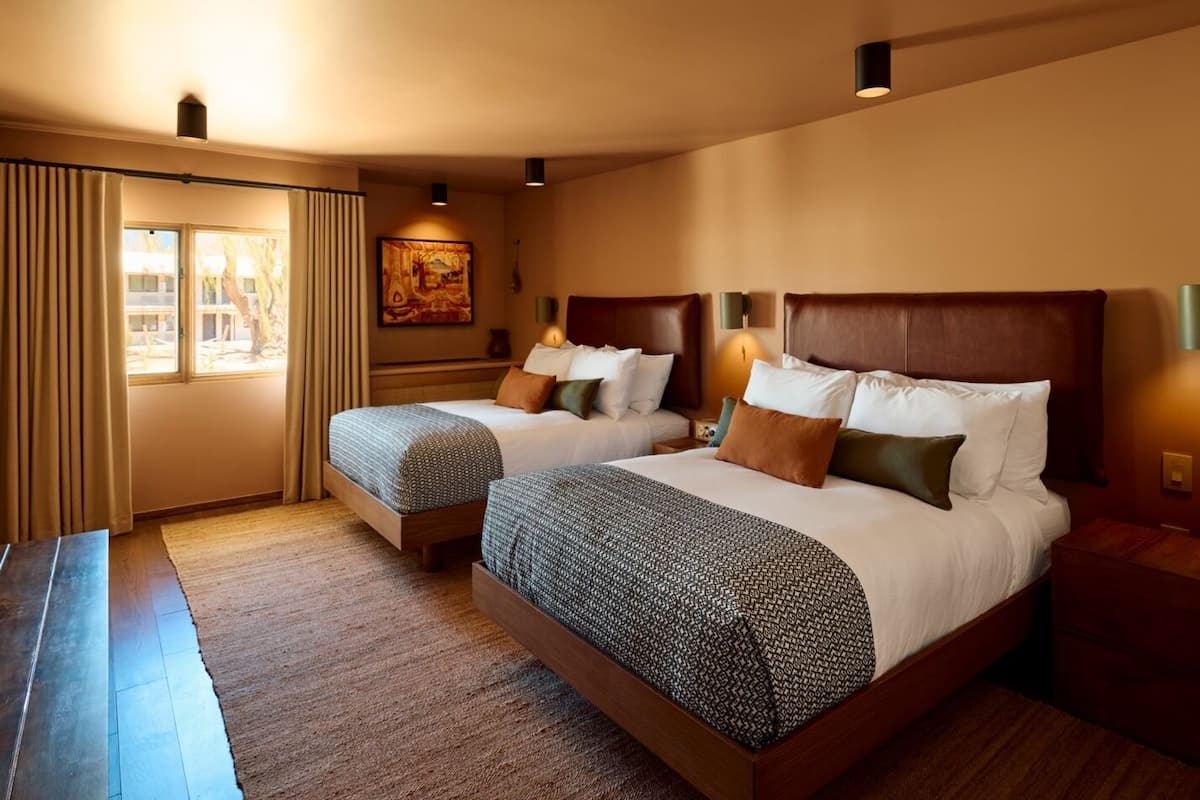 A hotel room with two beds, a large window with curtains, a painting on the wall, and a wooden floor.