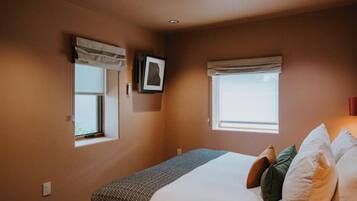 Un dormitorio con cama, dos ventanas con cortinas, un cuadro en la pared y una lámpara en el techo.