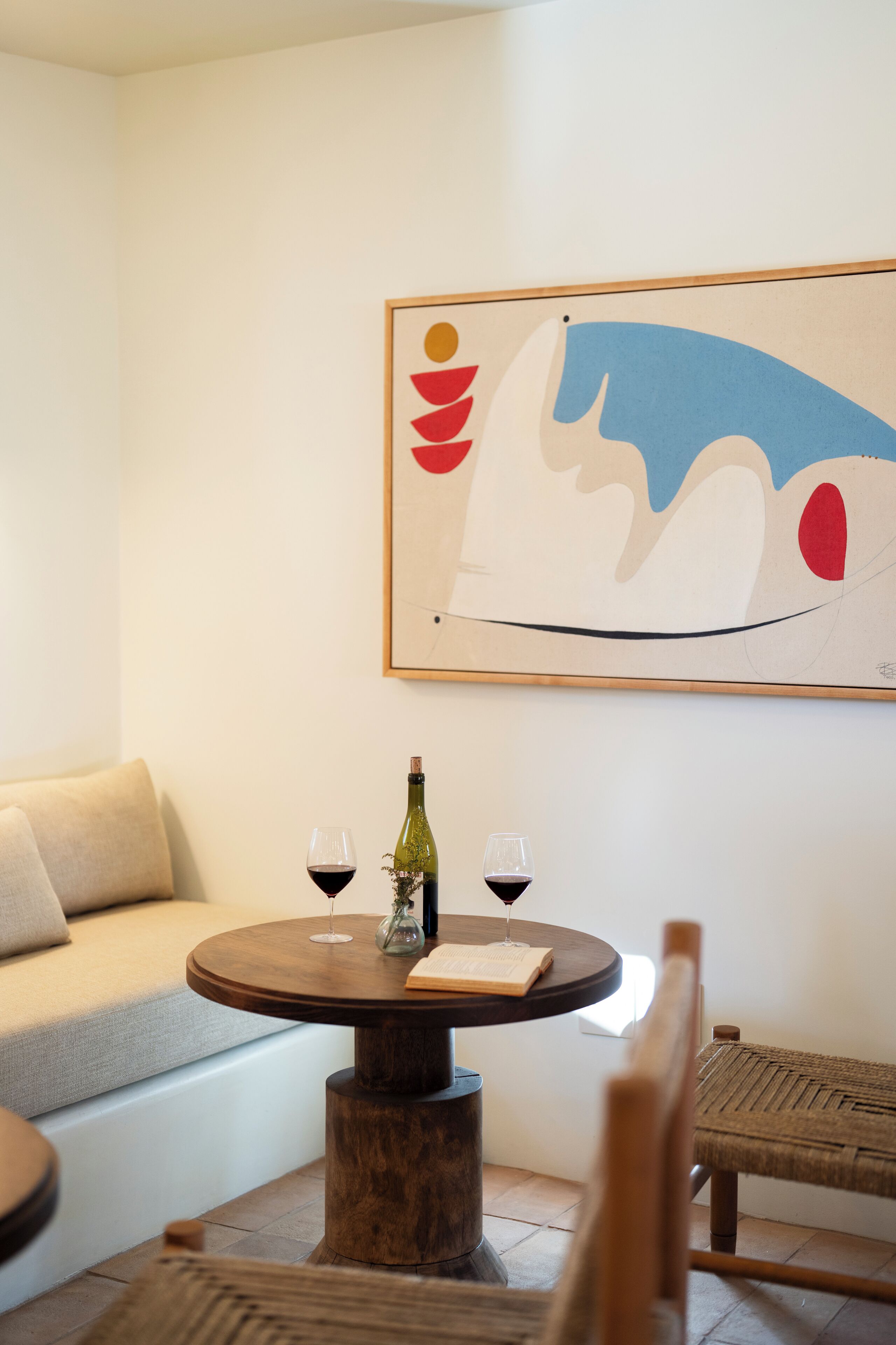 Un espacio de sala acogedor con una mesa redonda de madera, dos copas de vino y una botella. Un cuadro abstracto y colorido adorna la pared sobre la mesa.