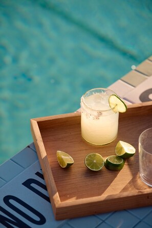 Un vaso de una bebida de color claro con una rodaja de lima sobre una bandeja de madera junto a una piscina.