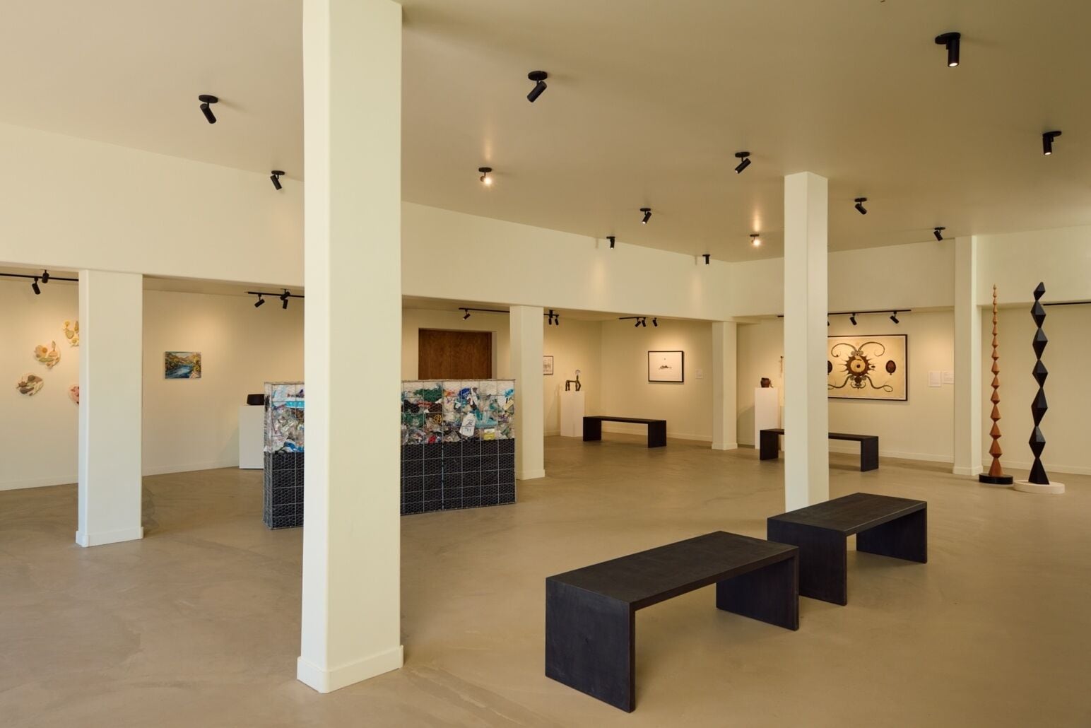 Eine Kunstgalerie mit weißen Säulen, schwarzen Bänken und verschiedenen Kunstwerken an den Wänden.