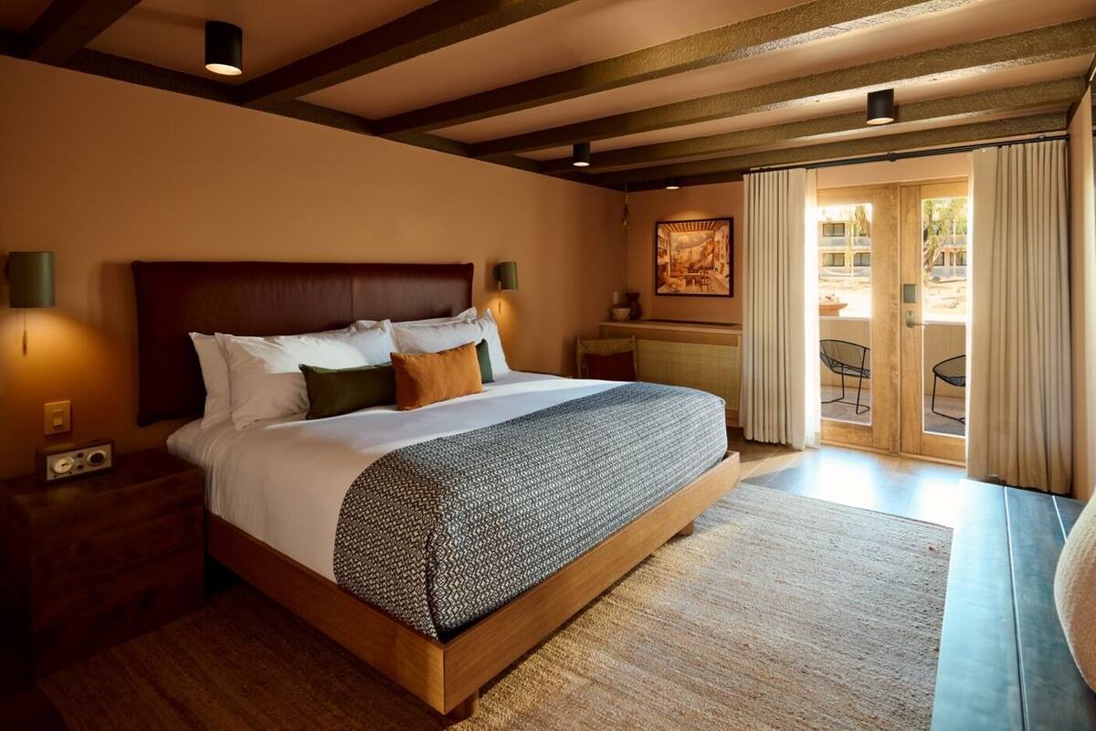A well-lit bedroom with a large bed, a nightstand, a desk with a chair, and a view of the outside through a glass door.