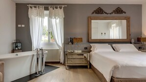Una habitación de hotel con bañera, un espejo grande, una mesita de noche con lámpara y una cama con ropa blanca.