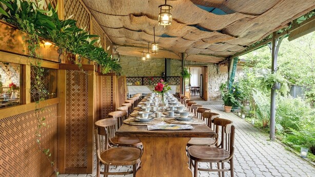 Una larga mesa de madera dispuesta para comer, con sillas, rodeada por un techo y paredes de madera rústica, con plantas y vistas al exterior.