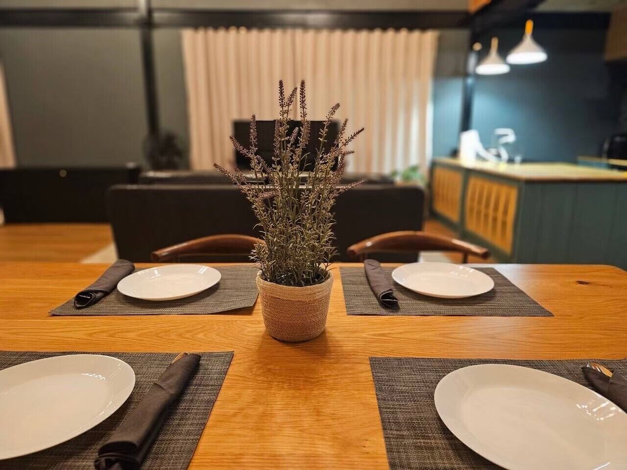 A dining table with four white plates, two dark napkins, and a woven pot with dried plants.