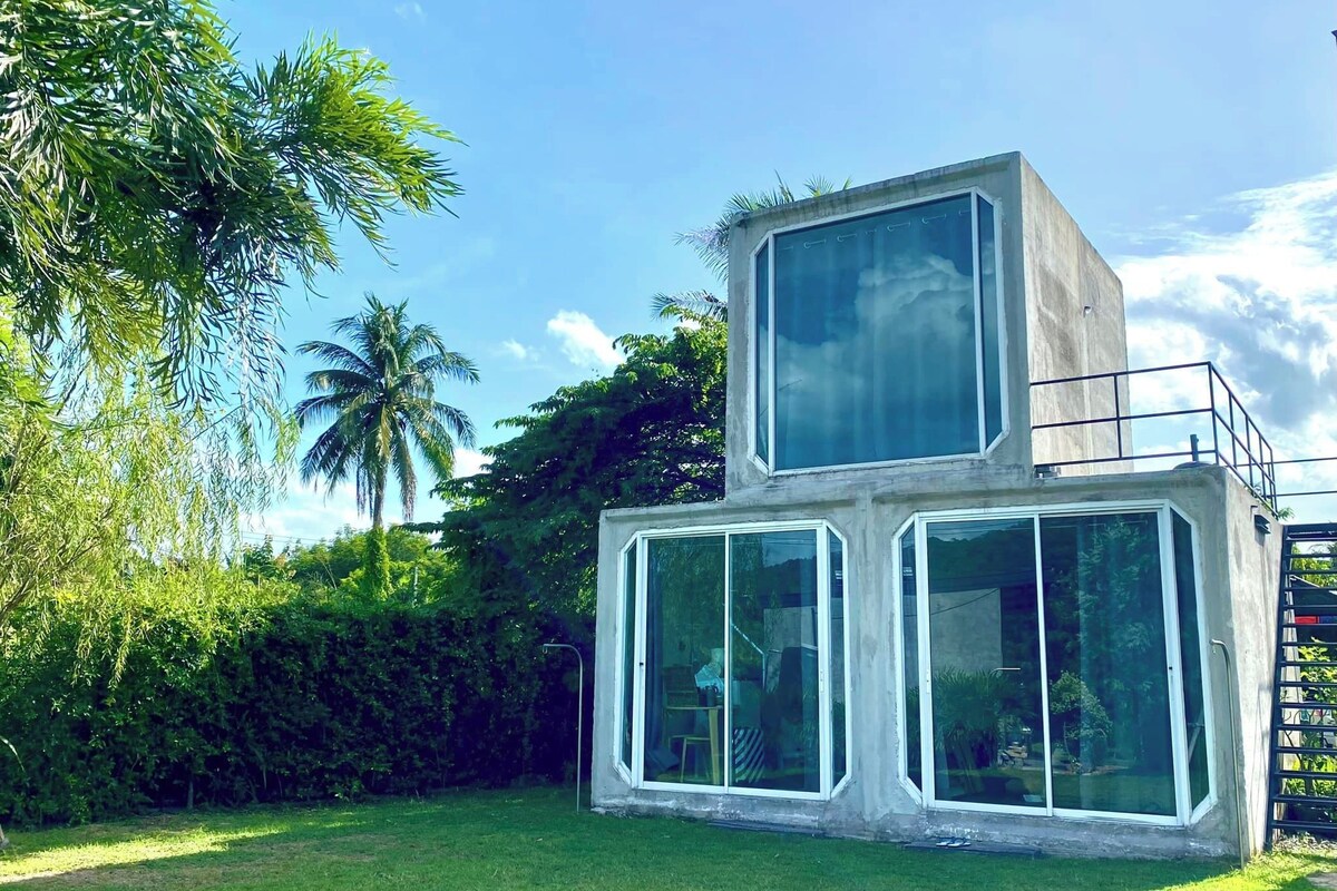 A modern, concrete building with large windows and a flat roof, surrounded by lush greenery and palm trees.