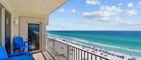 A balcony with blue chairs and a table, overlooking a beach with umbrellas and people.