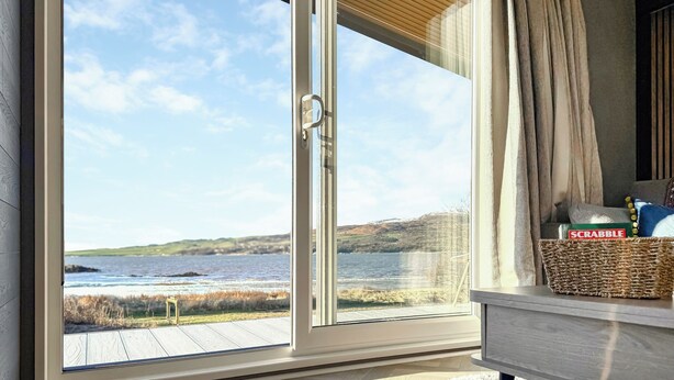 A room with a large sliding glass door leading to a view of a lake and hills. - 1 bedroom accommodation in Sandgreen (Sandgreen)