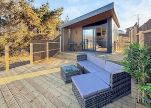 A modern outdoor deck with wicker furniture, a wooden fence, and a building with large glass doors. - 1 bedroom accommodation in Sandgreen (Sandgreen)