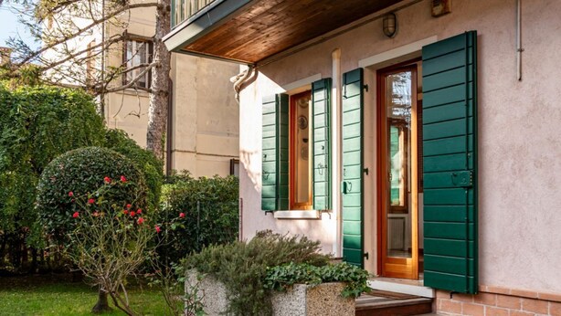 A building with a stone facade, green shutters, and a small garden with a bush and red flowers. - GuestHost - Cozy 2-Bedroom Stay at Lido di Venezia (Lido)