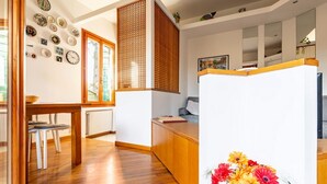 A room with a wooden floor, a dining table, a kitchen area, and a white partition with a floral arrangement. - GuestHost - Cozy 2-Bedroom Stay at Lido di Venezia (Lido)