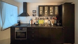 Une cuisine de taille réduite, dotée de meubles de rangement sombres, d’un four encastré et d’une fenêtre avec des stores.