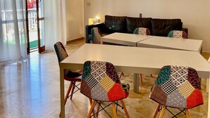 Un coin repas avec une table blanche, deux chaises aux motifs colorés, un canapé en cuir noir et une porte-fenêtre coulissante donnant sur l’extérieur.