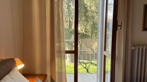 Une chambre avec un lit, une table de chevet, une lampe et une vue sur un espace extérieur verdoyant, accessible par une porte vitrée.