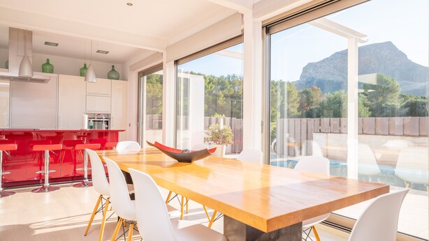 A modern kitchen with a wooden table and white chairs, large windows with a mountain view, and a red bar area. - VILLA MONTFARRUTX - Modern villa with private swimming pool and free Wifi (Artà)