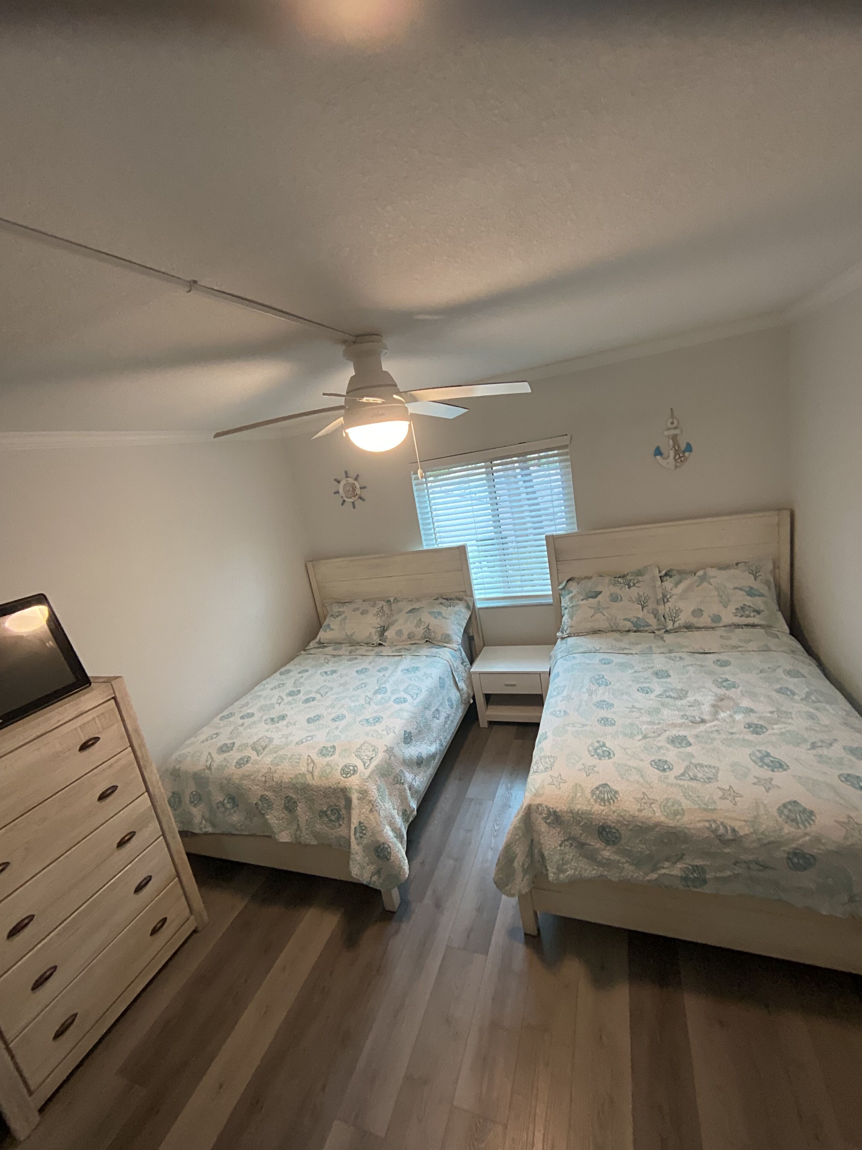 Une chambre avec deux lits, un ventilateur de plafond, une table de chevet, une commode et une fenêtre avec des stores.