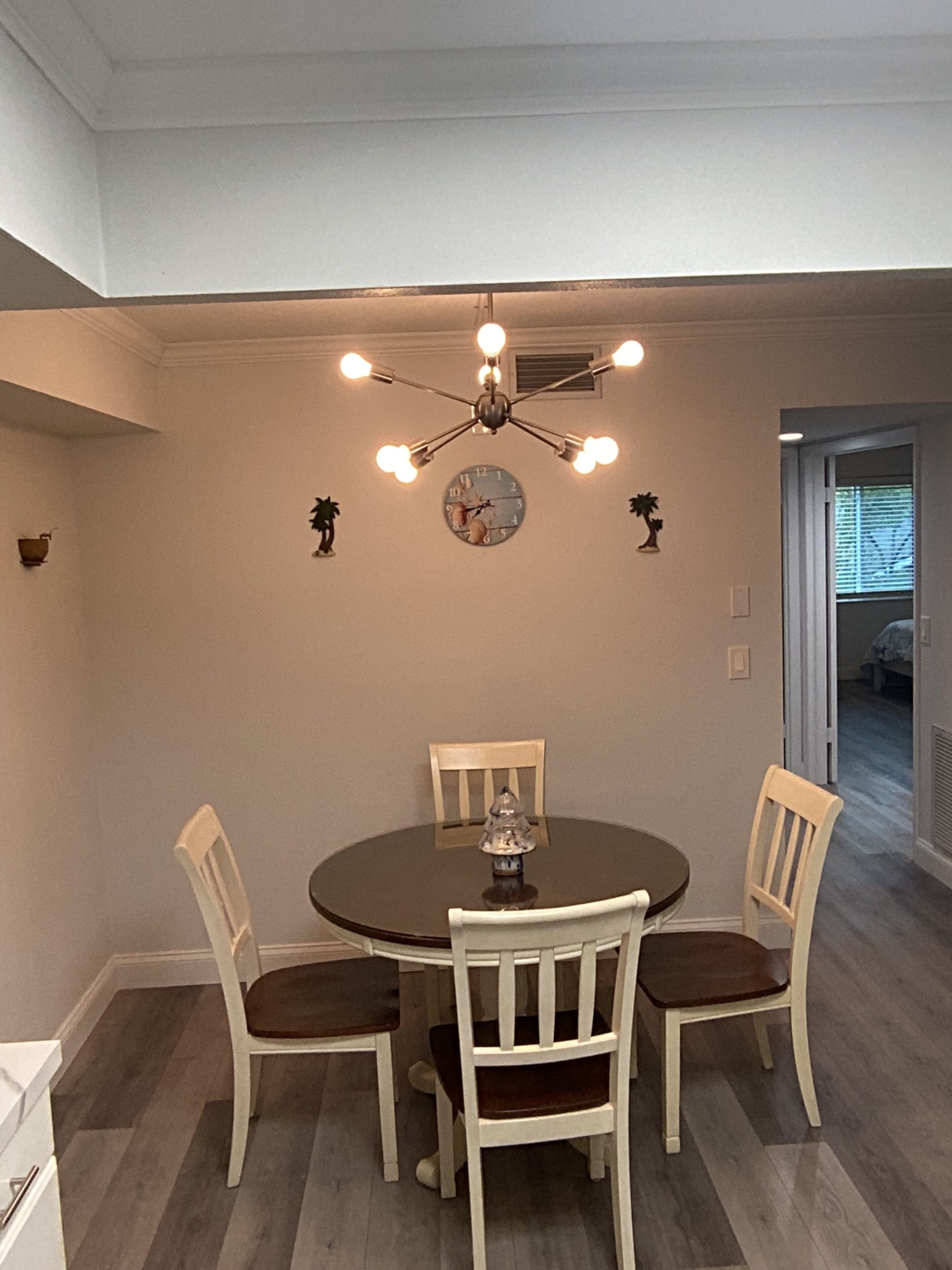 A dining area with a round table, four chairs, a chandelier, and a wall clock.