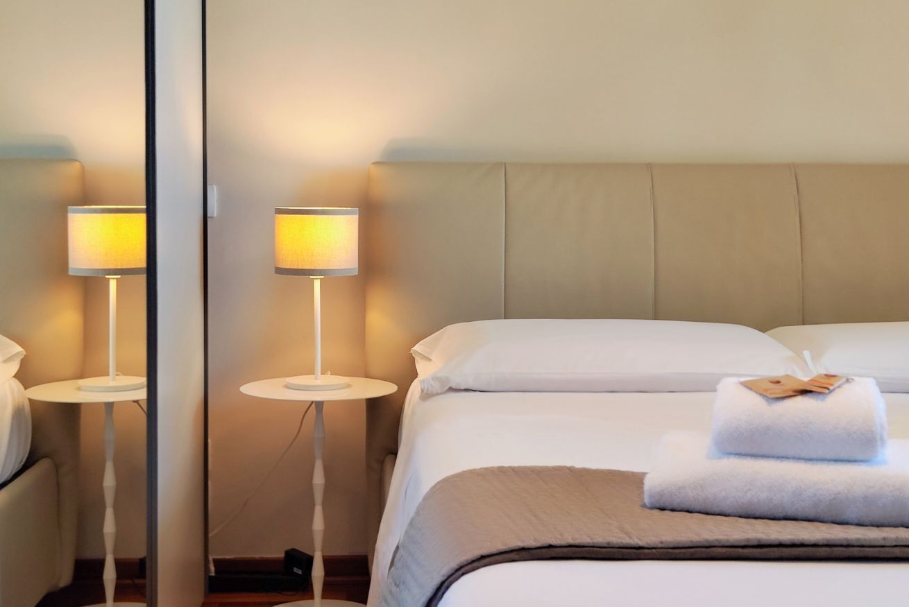 Et hotelværelse med en seng, to sengeborde med lamper, et spejl og en pænt redt seng med et håndklæde.