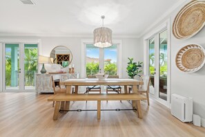 A spacious dining area with a wooden table and benches, large windows with a view of greenery, a hanging light fixture, and a decorative mirror. - Luxurious 3-bedroom home with Pool and Dock in Key Colony Beach (Key Colony Beach)