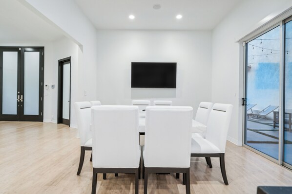 A modern dining area with a large table and white chairs, a flat-screen TV mounted on the wall, and a view of a balcony with outdoor furniture.