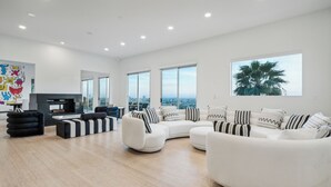 A spacious living room with a large white sectional sofa, a black and white striped ottoman, and a flat-screen TV. There are large windows offering a city view, and a large framed picture of a palm tree on the wall. - The Hollywood Hills Luxury Nest with Views, Pool and Spa. (Los Angeles)