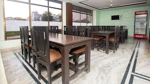 A dining area with multiple wooden tables and chairs, large windows with a view of buildings, a ceiling fan, and a Coca-Cola refrigerator.