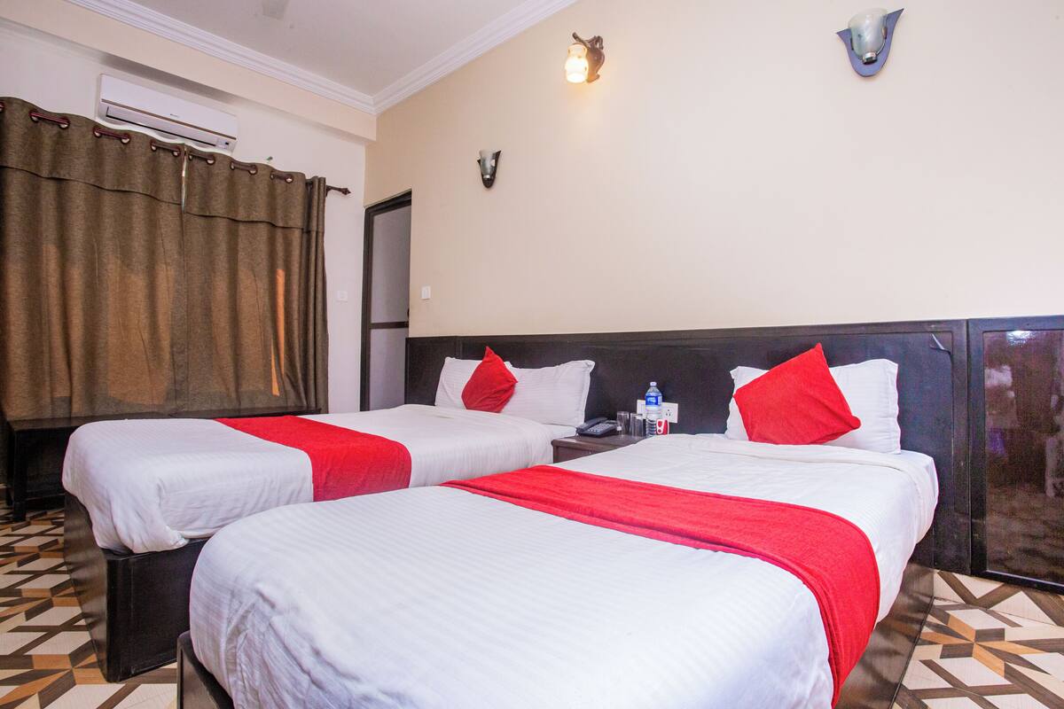 A hotel room with two beds, each with white bedding and red accents, a minibar, and a wall-mounted air conditioner.