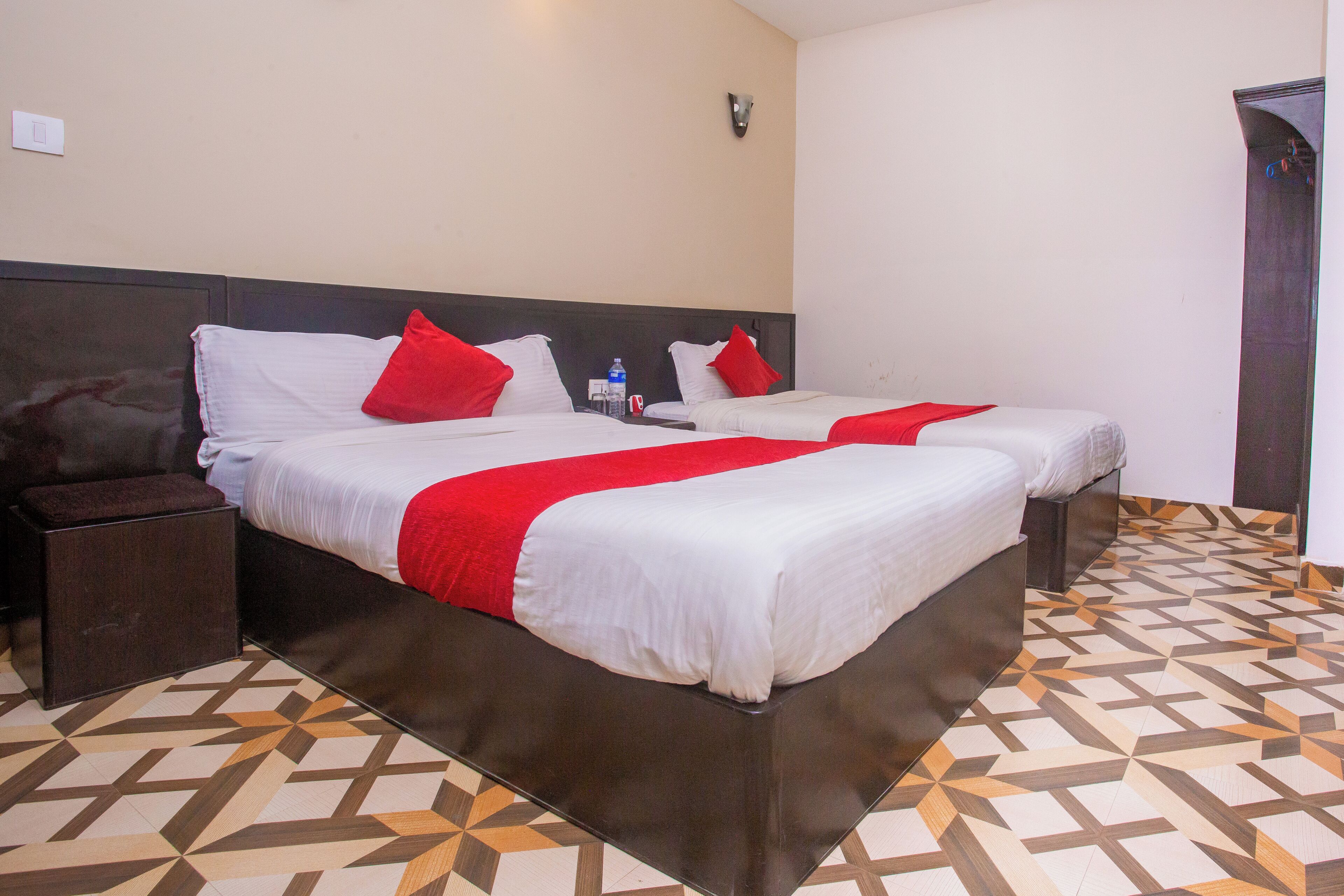 A hotel room with two beds, each with white linens and red pillows, a bedside table, and a patterned floor.