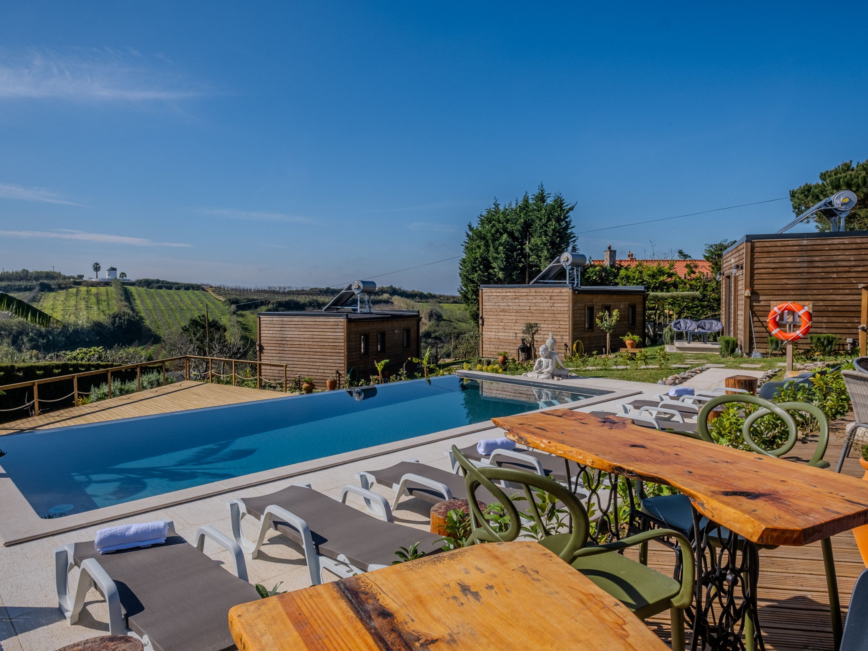 Zona de piscina con sillones de descanso y una mesa de madera, rodeada de cabañas de madera y vistas al campo.