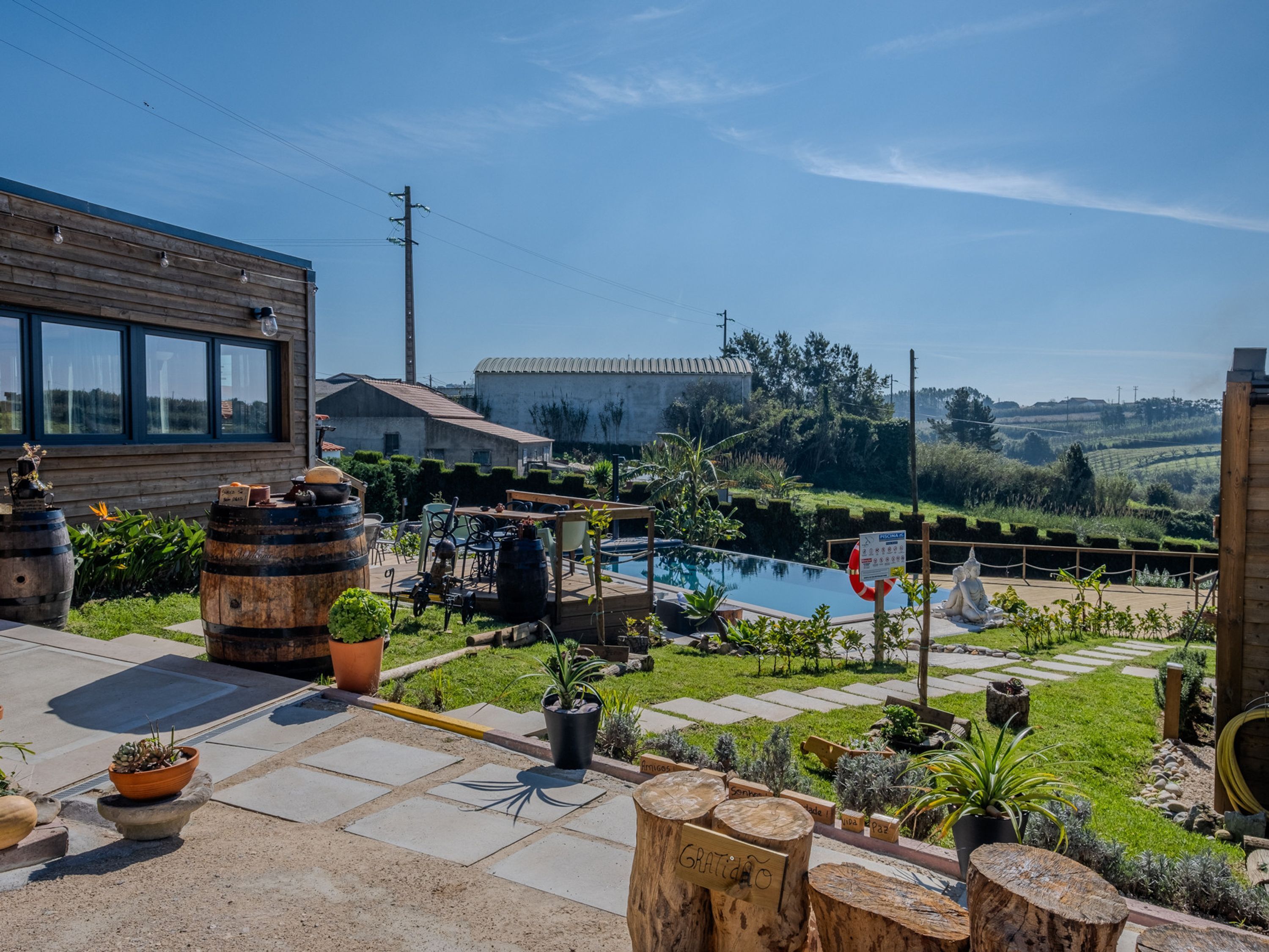 Zona de piscina con terraza de madera, plantas en macetas y vistas al campo.