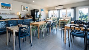 A spacious dining area with a long wooden table and chairs, a kitchen area with blue cabinets, and large windows offering a view of the outdoors. - Villas P Moinho - 04 Macieira (Obidos)