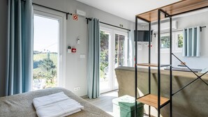 A modern hotel room with a large bed, a green ottoman, a wooden side table, and a balcony with a view of greenery. - Villas P Moinho - 04 Macieira (Obidos)