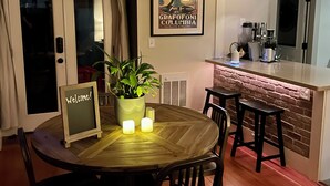 A dining area with a round wooden table, black chairs, a welcome sign, a potted plant, and a vintage poster on the wall.
