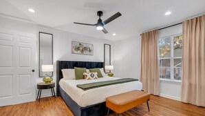 Une chambre à coucher comprenant un lit, des tables de chevet, un banc, un ventilateur de plafond et une fenêtre avec des rideaux.
