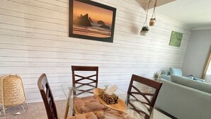 Een eetgedeelte met een glazen tafel, comfortabele stoelen, een houten stoel, een rieten mand, een lijst met een afbeelding en een bank op de achtergrond.