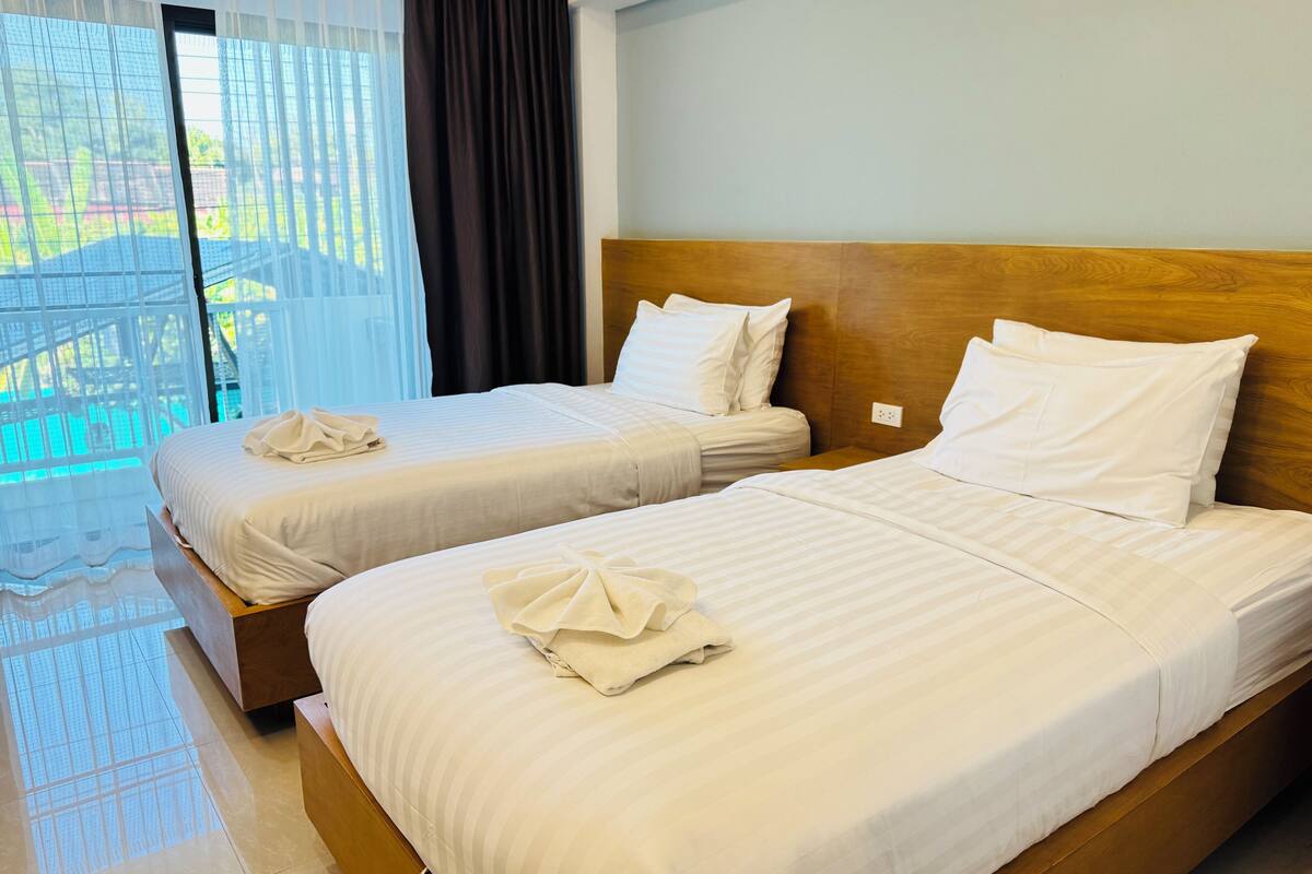 Habitación de hotel con dos camas, cada una con cabecero de madera, ropa de cama blanca y una toalla doblada con cuidado.