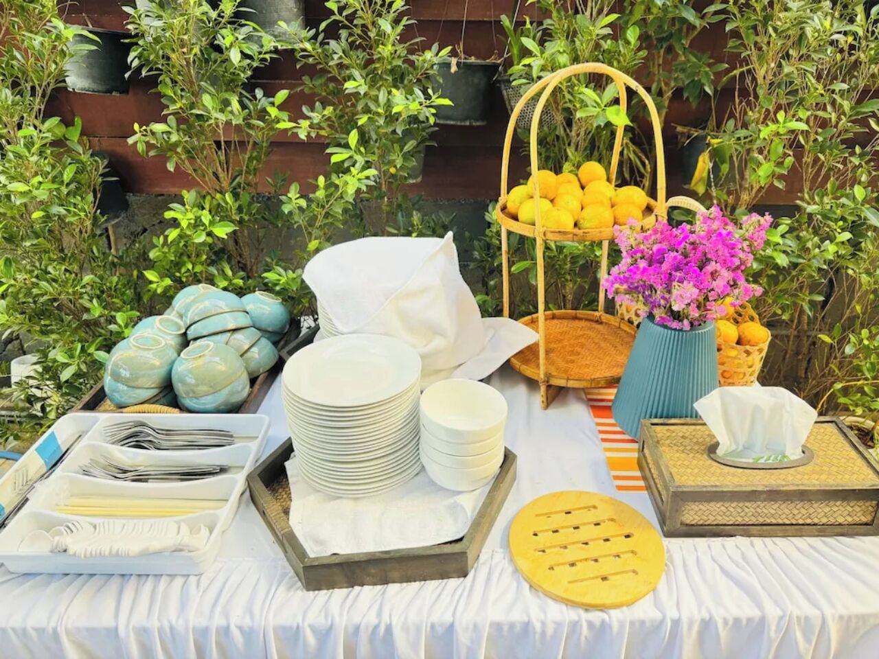 A table set with plates, bowls, and utensils, surrounded by potted plants and a basket of lemons.