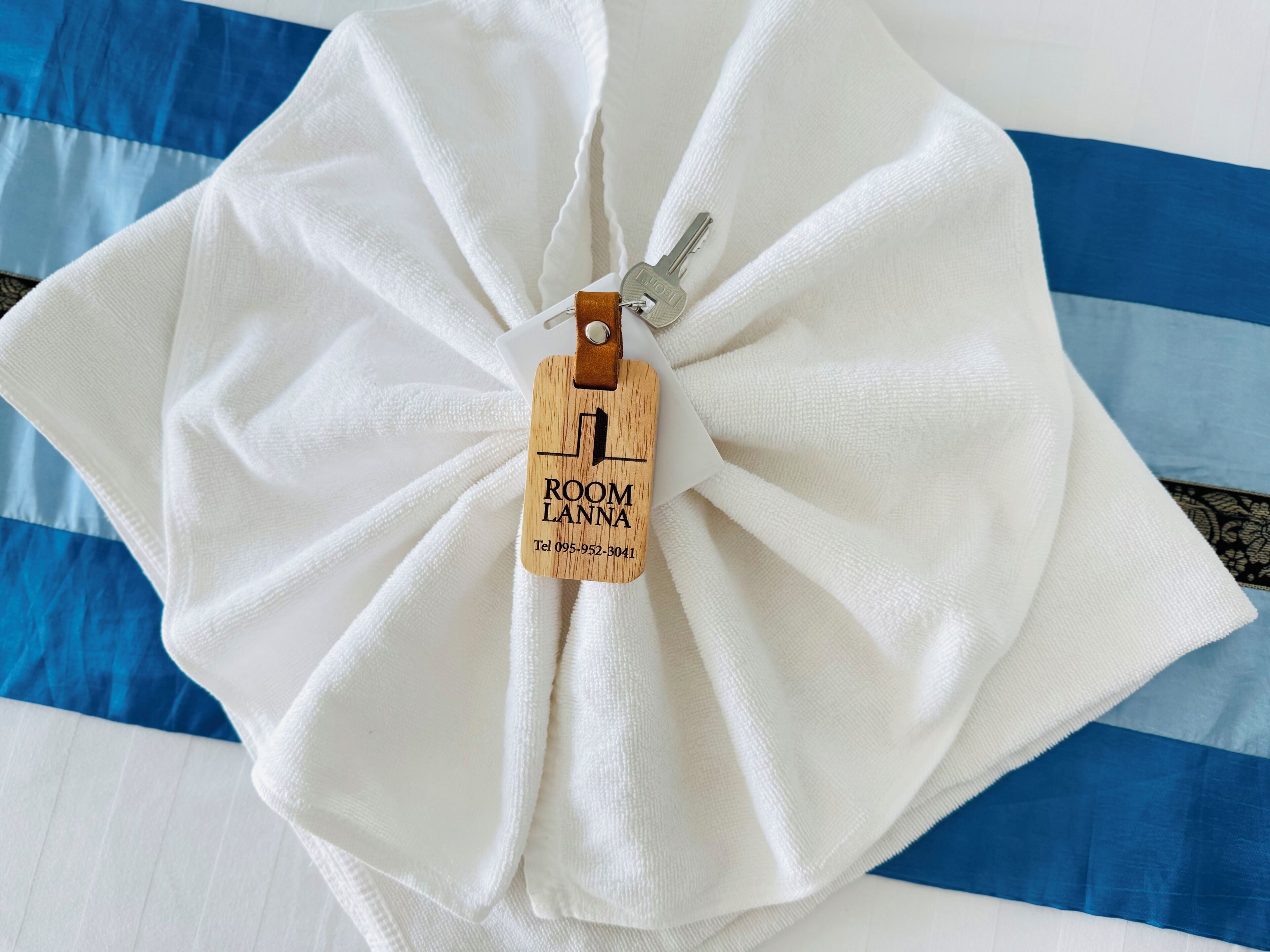 A neatly folded white towel with a wooden tag labeled "ROOM LANTA" and a contact number.