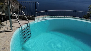 Ein klares, blaues Schwimmbecken mit Metallgeländer, umgeben von einer Steinterrasse mit Blick auf die Küste.