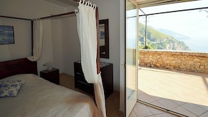 Ein Schlafzimmer mit Bett, Nachttisch, Kommode und einer Glastür, die auf einen Balkon mit Steinwand und Meerblick führt.