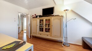 A room with a wooden floor, a white ceiling, a television on a cabinet, a floor lamp, and a bench.