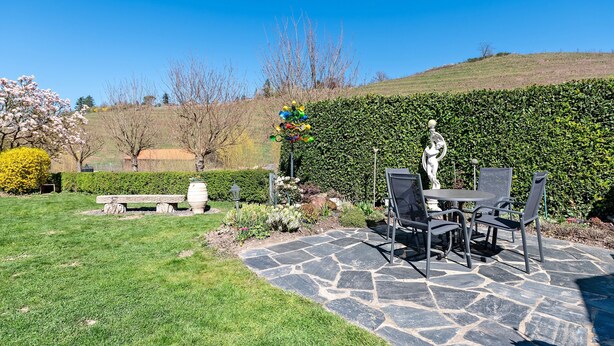 A patio with stone flooring, a table and chairs, a statue, and a hedge in the background.
