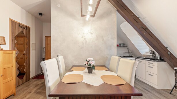 A dining area with a wooden table, white chairs, a vase with flowers, and a kitchen in the background.