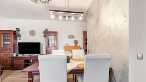 A living room with a wooden cabinet, a flat-screen TV, a dining table with chairs, and a ceiling light fixture.