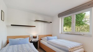 A bedroom with two single beds, a nightstand with a lamp, a window with a view of trees, and a wall-mounted shelf. - Apartment Knodel No. 3 in Kraichgau – Quiet, Wi-Fi & Ideal for Business (Sachsenheim)