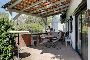 Eine überdachte Terrasse mit Tisch und Stühlen, eine Ziegelwand und Blick auf ein Haus.