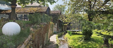 A narrow path leading to a rustic building with a thatched roof, surrounded by greenery and a small stream.
